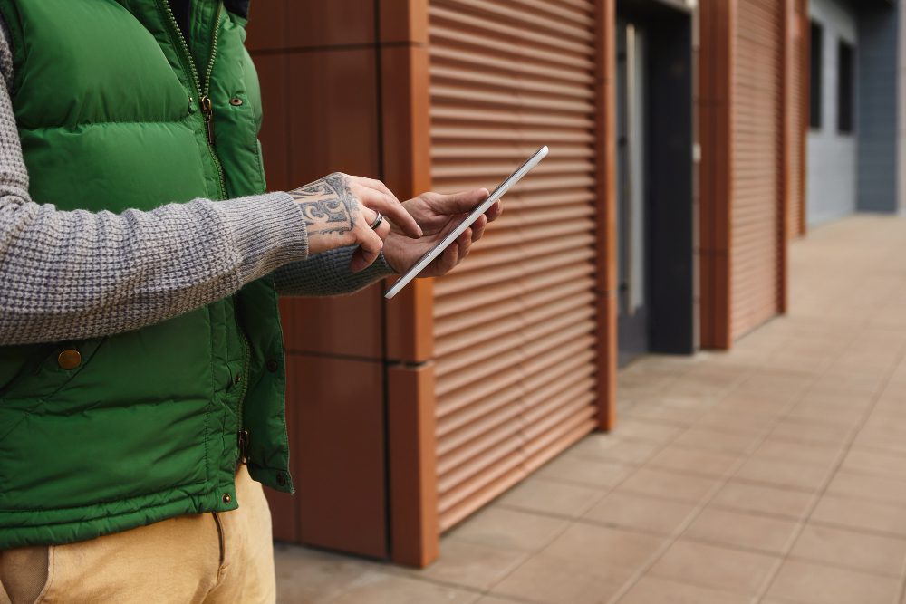 Person in a green puffer vest and gray knit sweater types on a tablet outdoors beside a building with wooden paneling, tattoo visible on the hand.
