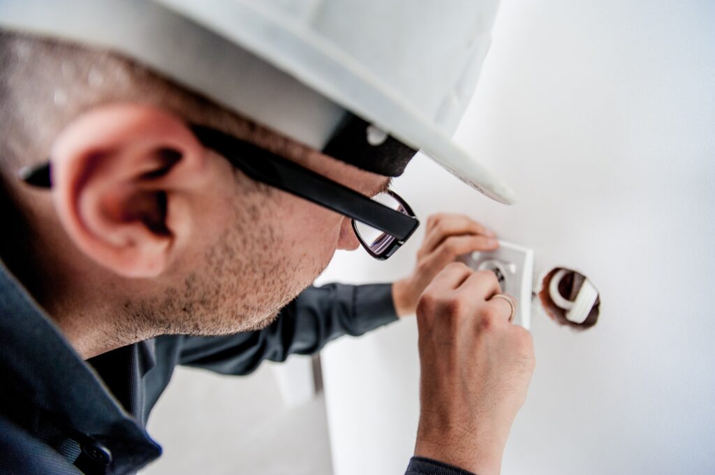 Electrician wearing a hard hat and glasses wiring an electrical outlet in a wall.