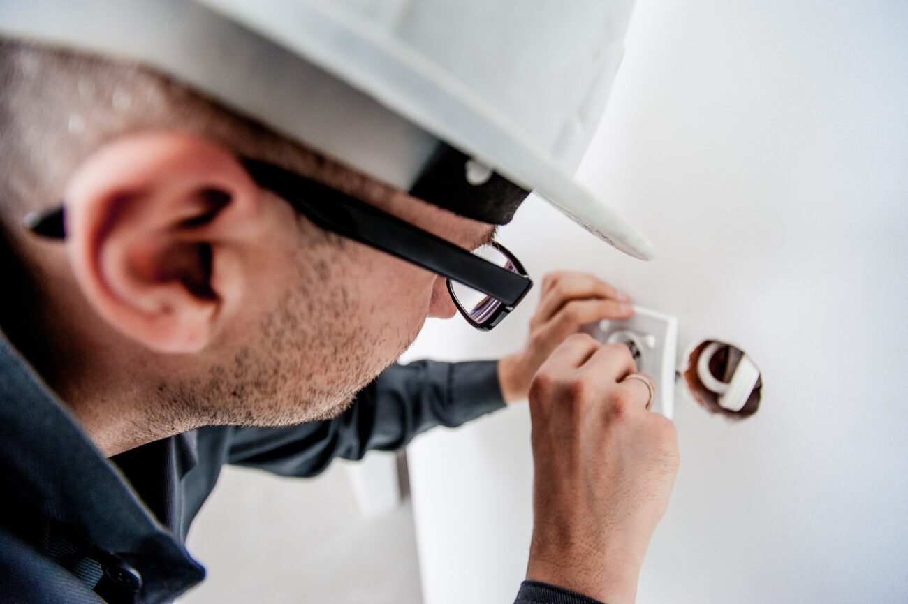 Electrician wearing a hard hat and glasses wiring an electrical outlet in a wall.