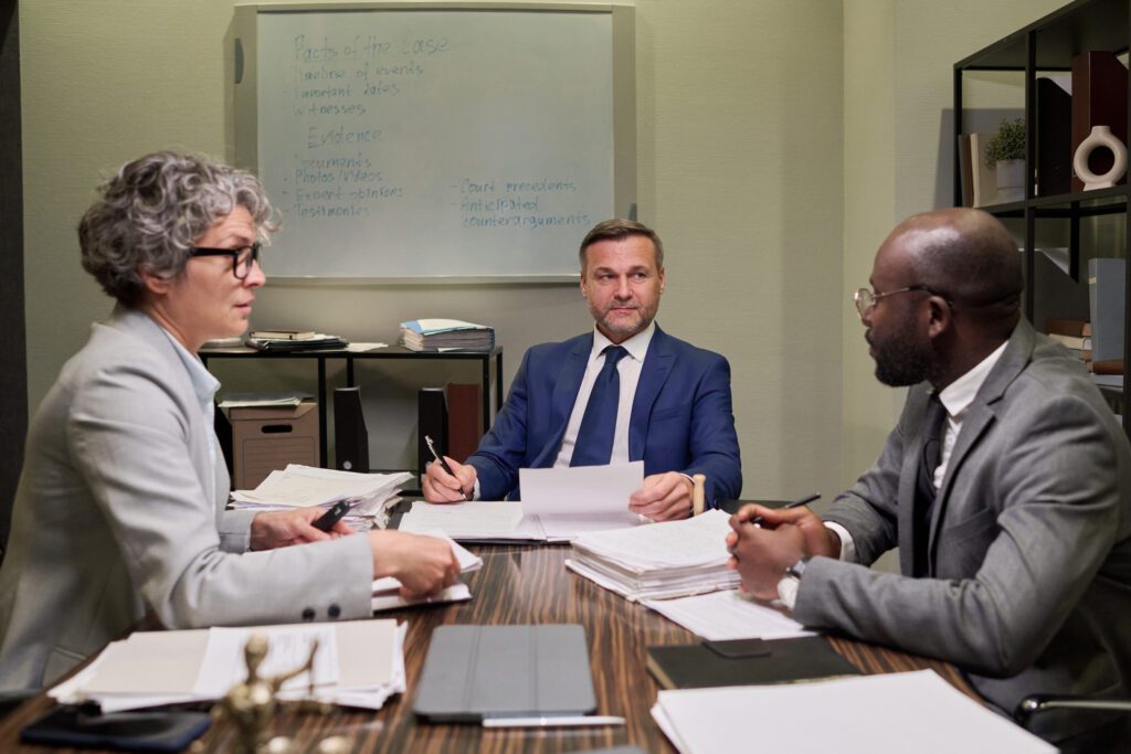 Three professionals in business attire sit around a wooden table with papers, discussing documents in a conference room.