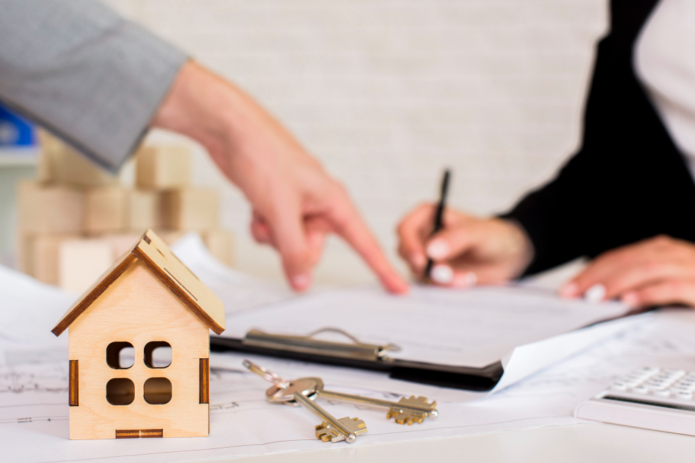 Small wooden house model on a desk with keys and a clipboard as two people sign documents in the background.