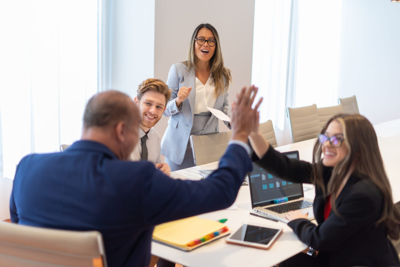 Office employee having a meeting with their team - Madrid Noticias 24h Team in a bright conference room celebrate with a high-five around a table with laptops and notebooks nearby.
