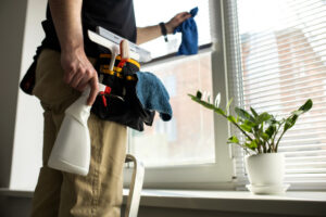 Person wearing a tool belt cleans a window, holding a spray bottle and a cleaning cloth in front of a sunlit window.