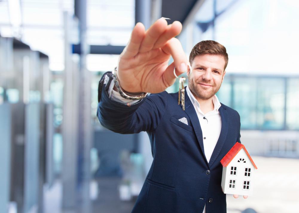 Smiling real estate agent in a navy suit holds up keys with a small white house model in a modern office setting.