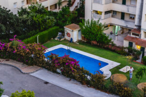 Residential courtyard with a blue rectangular pool, surrounded by flowering bushes and trees, and a person in the water nearby.