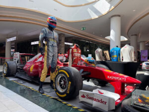 Red Formula 1 car on display in a mall, with a life-size racing suit and helmeted mannequin standing beside it, sponsor logos visible (Kaspersky, Santander).