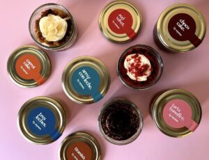 Top-down view of assorted dessert jars with color-coded labels on a pink surface, flavors include red velvet, carrot cake, tiramisu, arroz con leche, and chocolate cake.