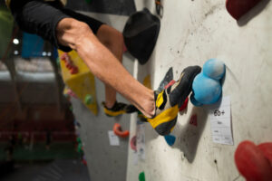 Climber's foot in a yellow and black climbing shoe resting on a blue hold on an indoor climbing wall; other holds and gym visible in the background.