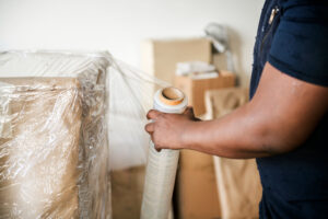 Person in a navy shirt seals a cardboard pallet with plastic wrap in a storage area: hands near a wrapped pallet with tape roll in hand.