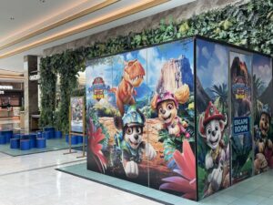 Colorful wall panels featuring Paw Patrol characters in a jungle scene inside a shopping mall; an escape room sign visible on the panels.