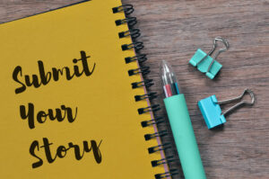Yellow spiral notebook with the words 'Submit Your Story' on a wooden desk, alongside a teal pen and binder clips.