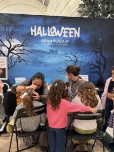 Children sit in chairs while adults apply Halloween makeup against a blue backdrop that says HALLOWEEN GRAN PLAZA 2° and features barren trees.