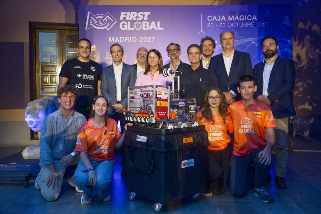 Group of students and organizers posing with a modular competition robot on a wheeled cart at the FIRST Global Madrid 2027 event.